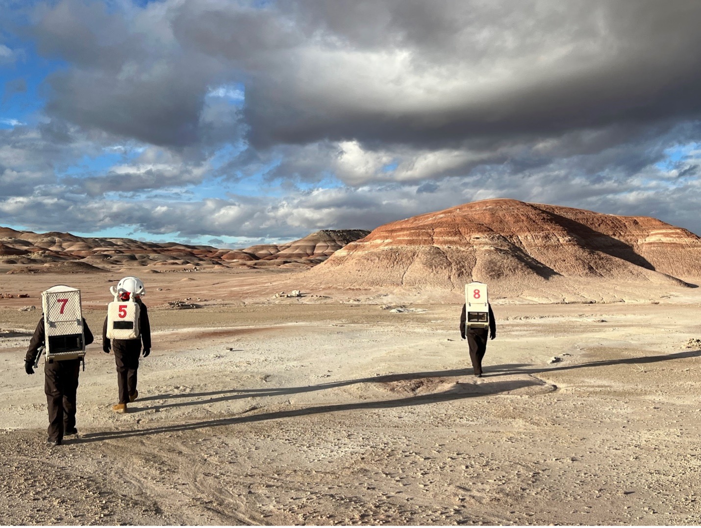 a view of a rock feature in the desert. three students in space suits walk in the foreground
