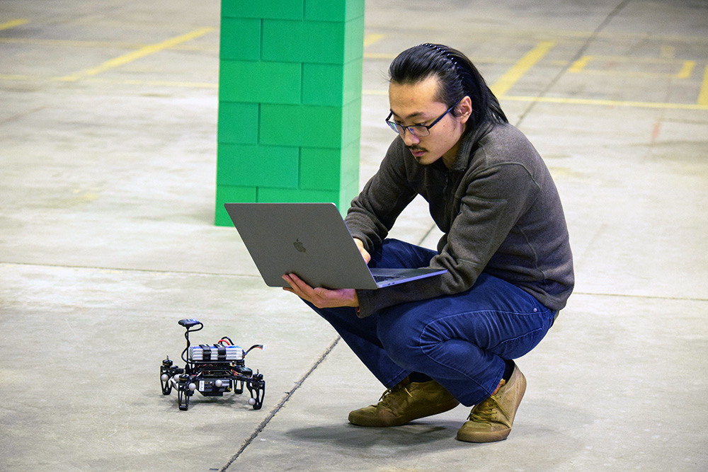 A student holding a laptop and squatting on a concrete floor next to a drone.