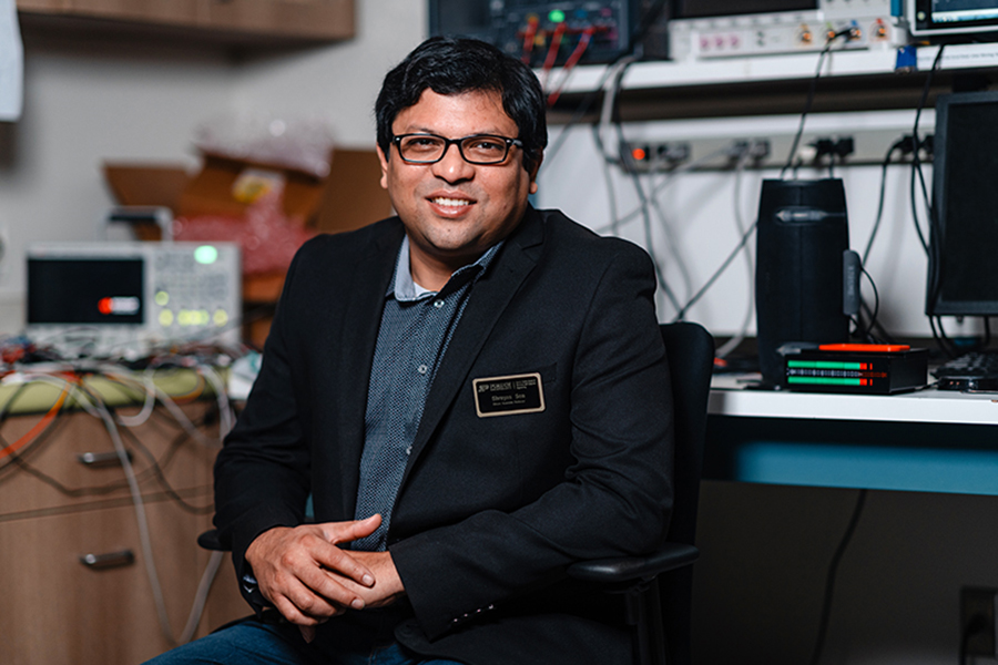 Professor Sen, dressed in a suit jacket and sweater, sits in front of various lab equipment.