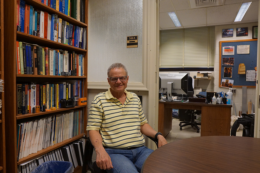 Professor Mike Melloch, dressed in jeans and a yellow striped polo, sits at a table just outside his office in the electrical engineering building. Behind his is a bookshelf and a view into his office where there is a desk and bulletin board.