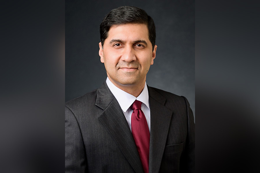 Vijay poses for a portrait. The background is nearly black. He is wearing a dark suit, white shirt, and red tie.