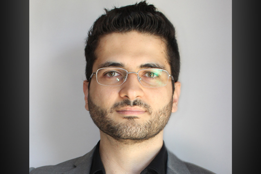 Portrait of Vincius Cabral do Nascimento. He is standing in front of a white backdrop, wearing a gray jacket, black shirt, and glasses.