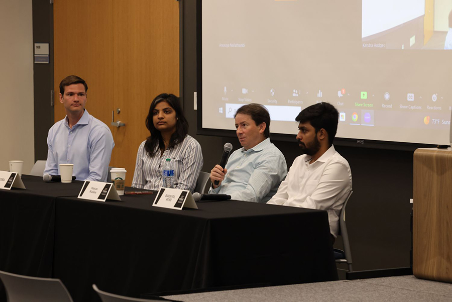 Three graduate students in the project track master's program sit beside Marcus Weldon during a panel discussion. They are seated at a table with a projector screen behind them.