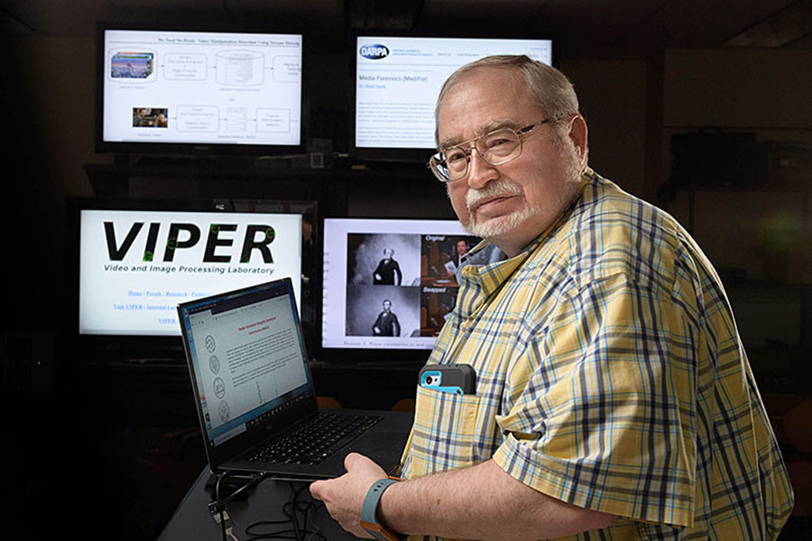 A man in a yellow and blue plaid shirt posing in front of several computers