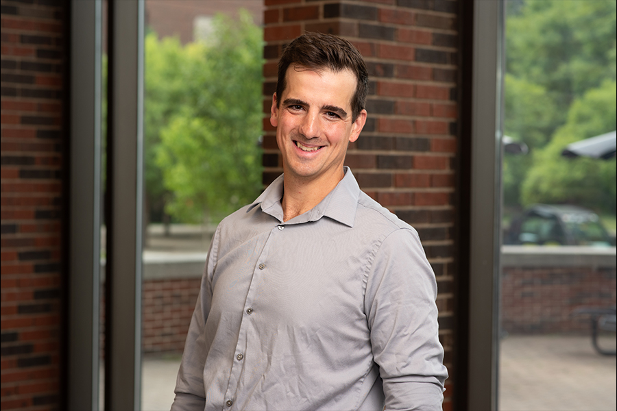 A man in a gray shirt stands confidently in front of a textured brick wall, smiling at the camera