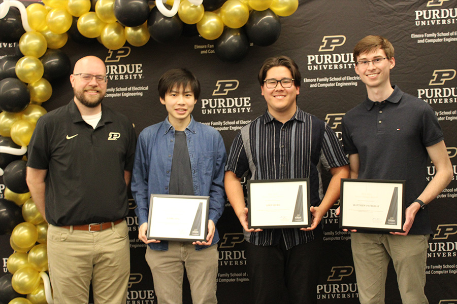 A group of four men holds their awards, smiling in front of a backdrop of vibrant balloons at a recognition ceremony
