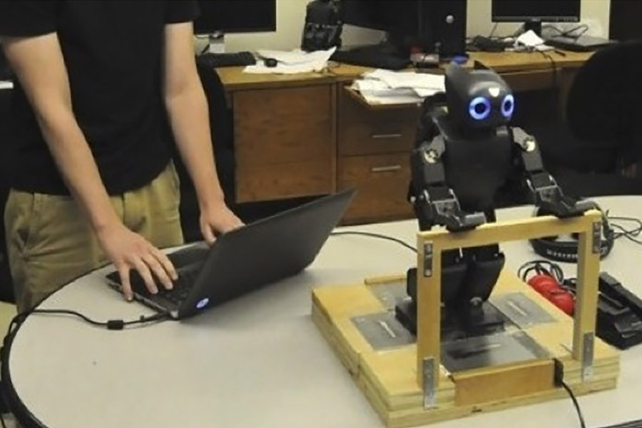 Man out of frame typing on laptop beside a small black robot on a wooden platform with wires connected.