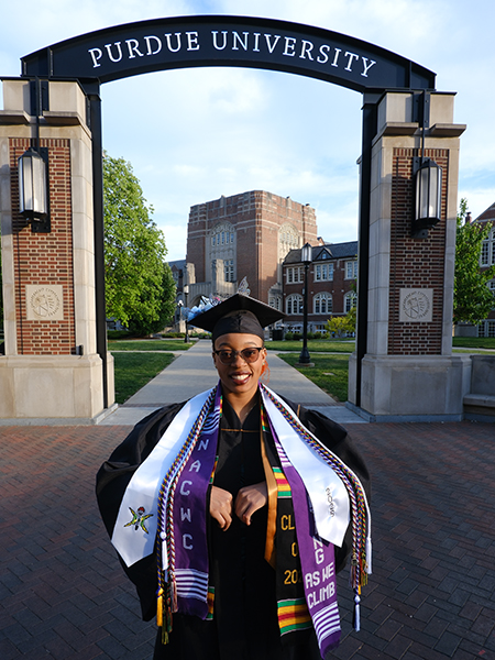 Student in gap and gown standing in front of archway that reads "Purdue University"