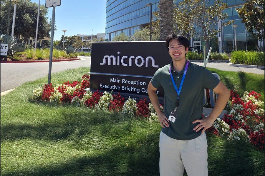 Student with dark hair and glasses smiling in front of sign that says "Micron"