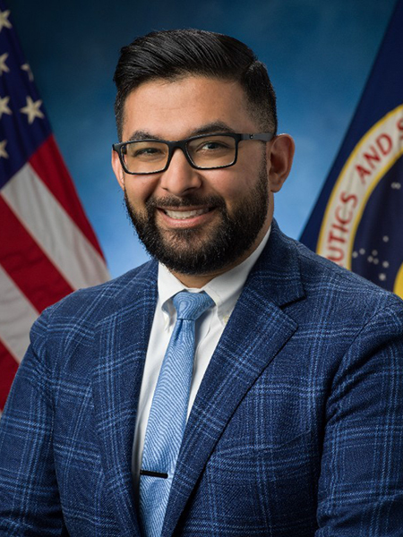 Man with dark hair and glasses, smiling, wearing suit and tie