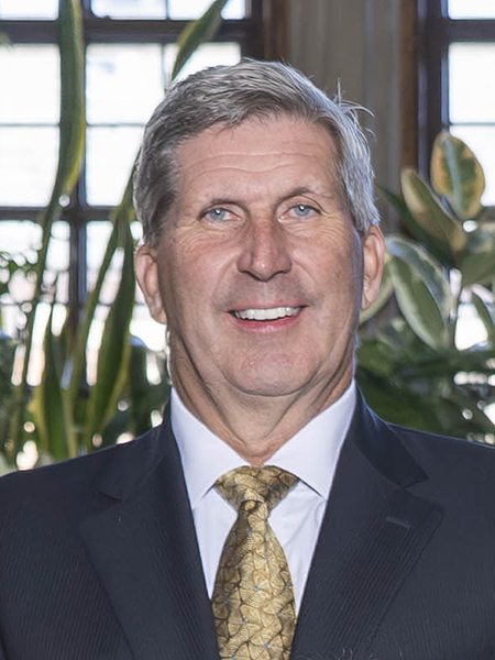 Man with gray hair and blue eyes, smiling, wearing a dark suit and gold tie