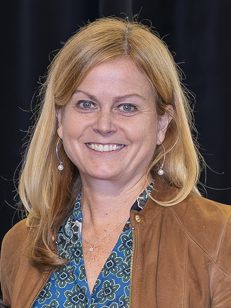 Woman with blonde hair and blue eyes, smiling, wearing blue patterned shirt and tan jacket