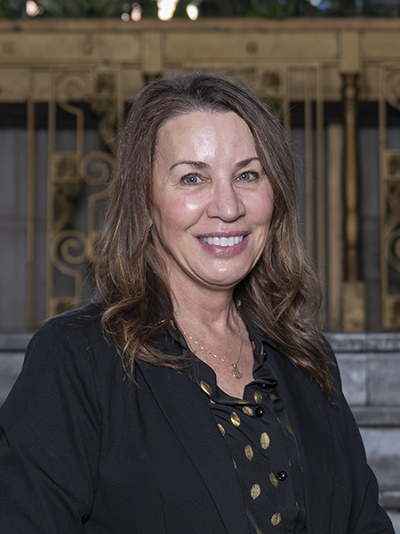 Woman with long brown hair and blue eyes, smiling, wearing dark jacket and polka dot scarf