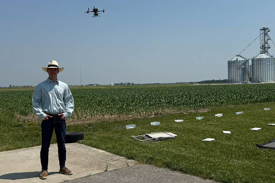 Student wearing hat, standing near cornfield with drone in air