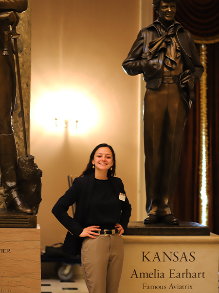 Student (Morton) with long dark hair smiling and posing next to a statue of Amelia Earhart. The statue plaque states "KANSAS Amelia Earhart Famous Aviatrix"