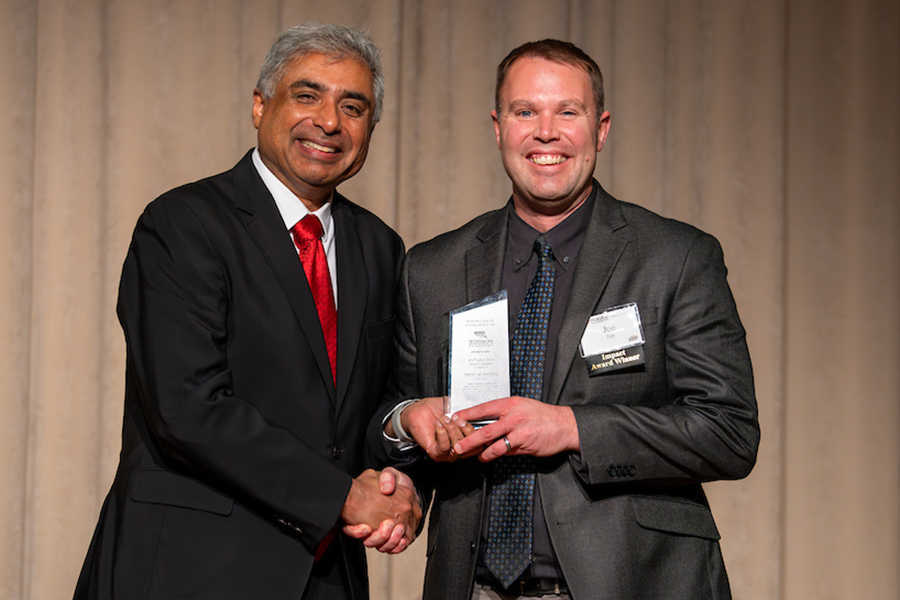 Two men standing next to each other, shaking hands, wearing suits, smiling