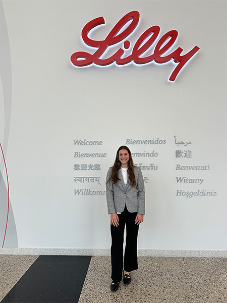 Student with long dark hair wearing a gray blazer and black pants, standing in front of a wall with "Lilly" in red 